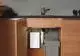 Weißer E-Kompaktdurchlauferhitzer unter der Küchenspüle, montiert an der Wand, mit angeschlossenen Wasserleitungen und einem Abfallbehälter daneben. Produkt von CLAGE.